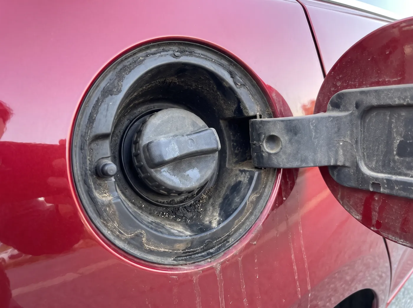 Geöffnete Tankklappe eines roten Autos, der Rand ist schwarz verkrustet, Deckel und Kunststoff sind staubig.